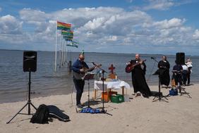 Am Strand von Dangast ist ein Altar aufgebaut. Zwei Personen spielen Gitarre. Weiße Wolken am blauen Himmel. Die Regenbogen-Fahnen wehen im Wind.